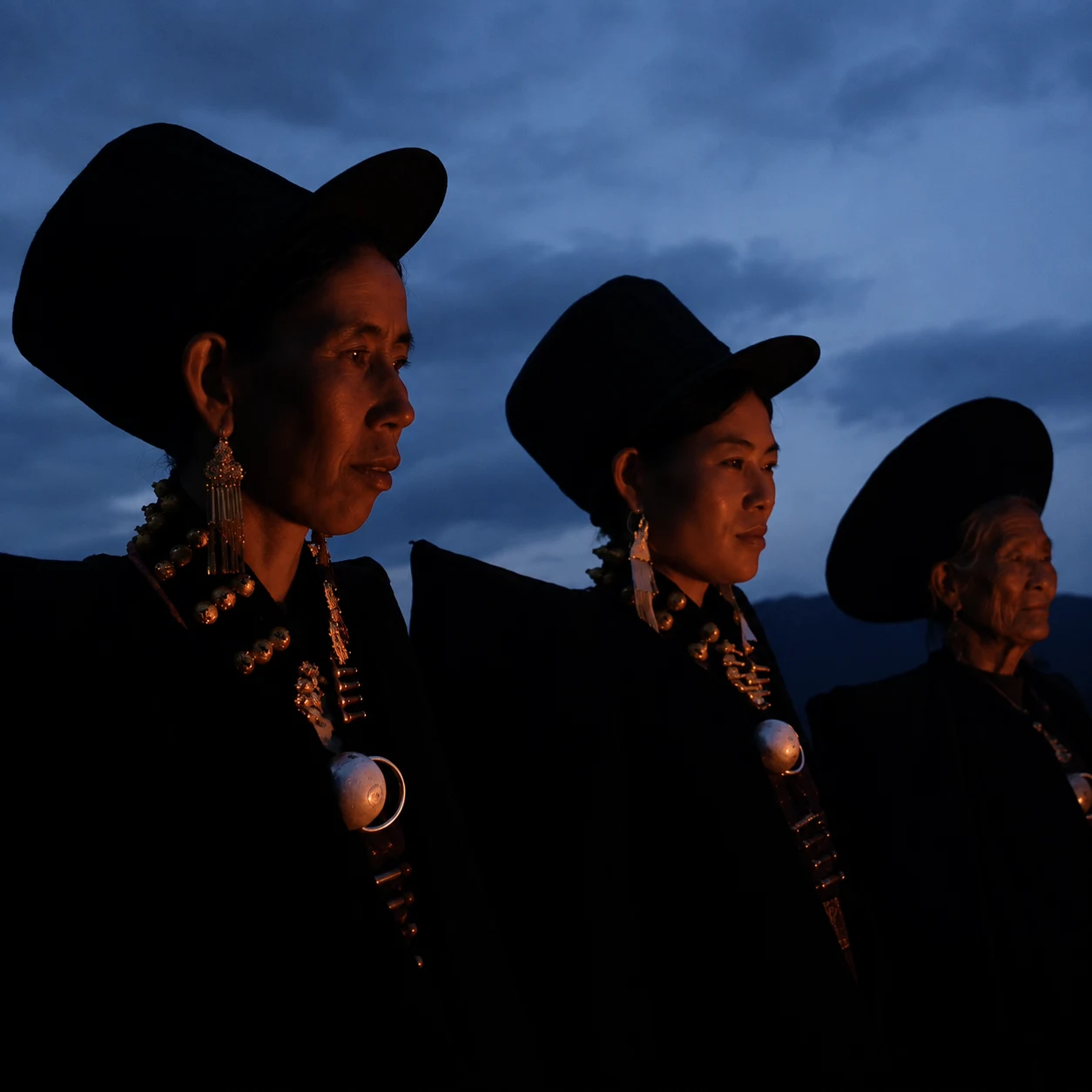 Three Yi women standing in ceremonial firelight, wearing traditional dress and silver ornaments
